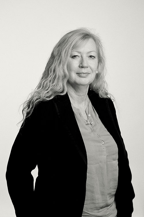 Frau mit langen, lockigen Haaren in einem schwarzen Blazer, neutraler Hintergrund.
